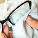 a person wiping the lenses of a pair of spectacles with a blue microfiber cloth while holding the glasses in one hand.