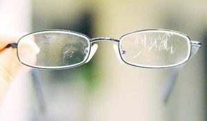 A person holding a pair of eyeglasses up to a bright light, with their eyes examining the lenses for any scratches or damage.